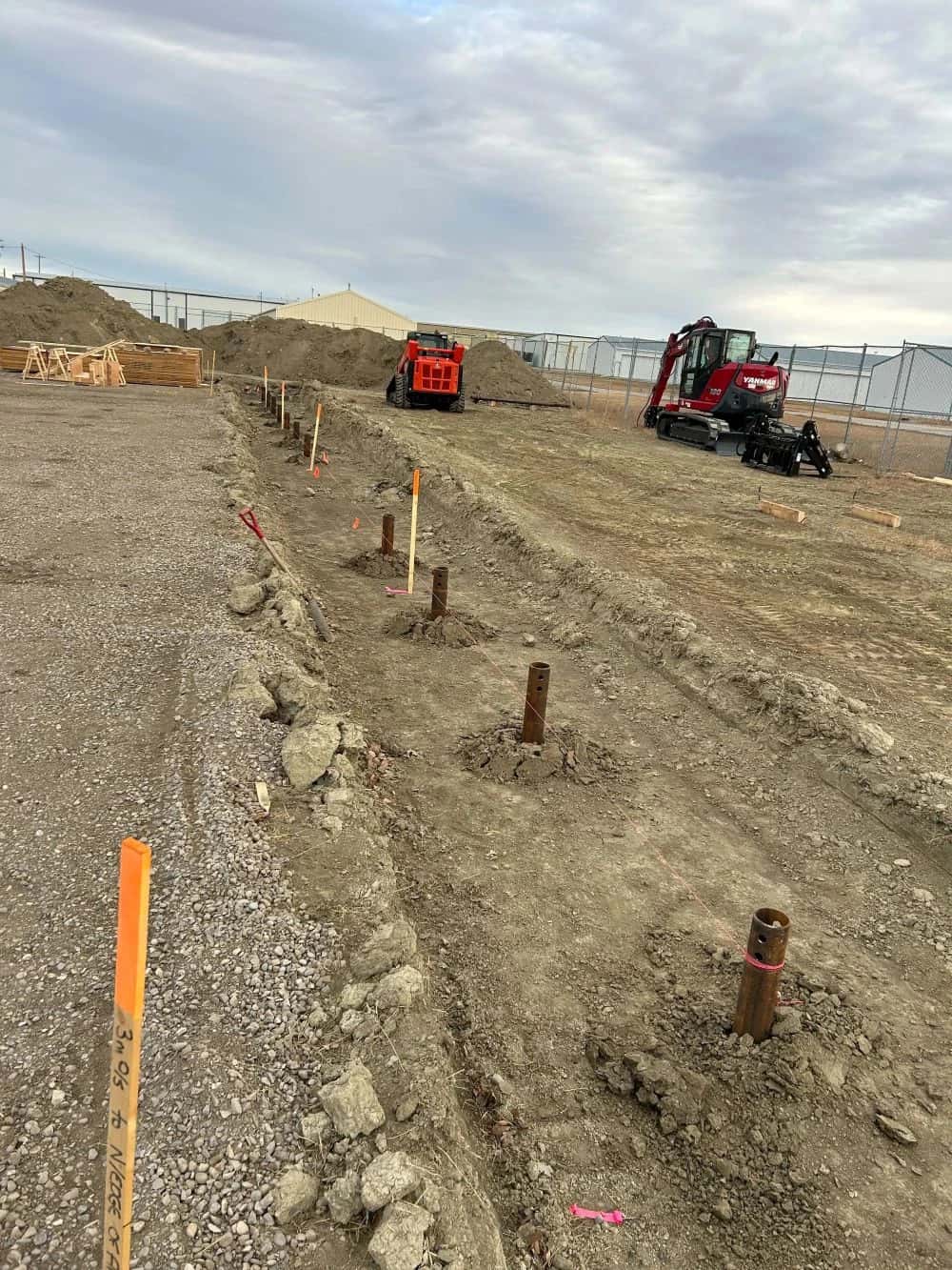 Screw-pile foundation layout staked out on a graded commercial site with an excavator and skid steer