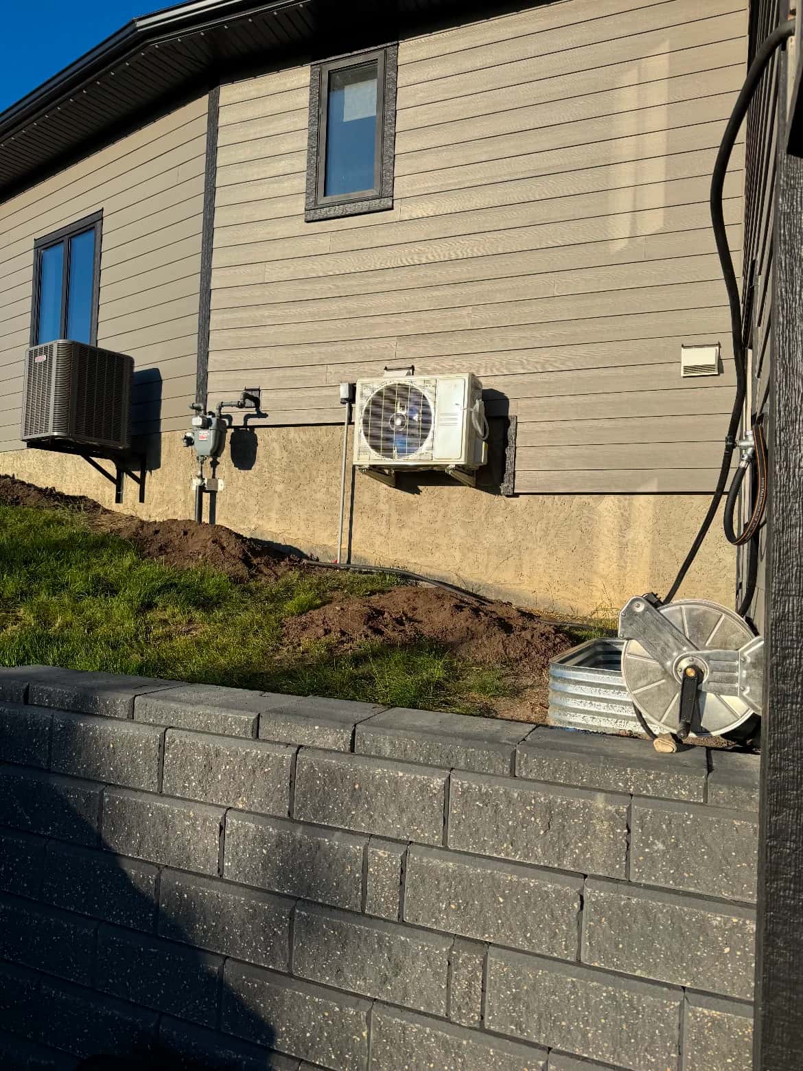 Residential exterior with air conditioner, mini-split condenser and gas meter above a block retaining wall